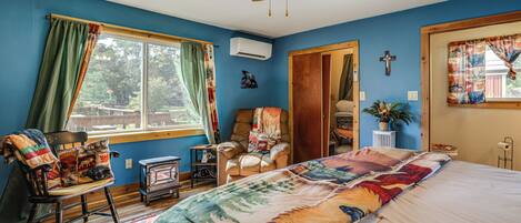 Ein Schlafzimmer mit einem großen Bett, einem Sessel mit Decke, Holzboden, Deckenventilator und einem Fenster mit Vorhängen.