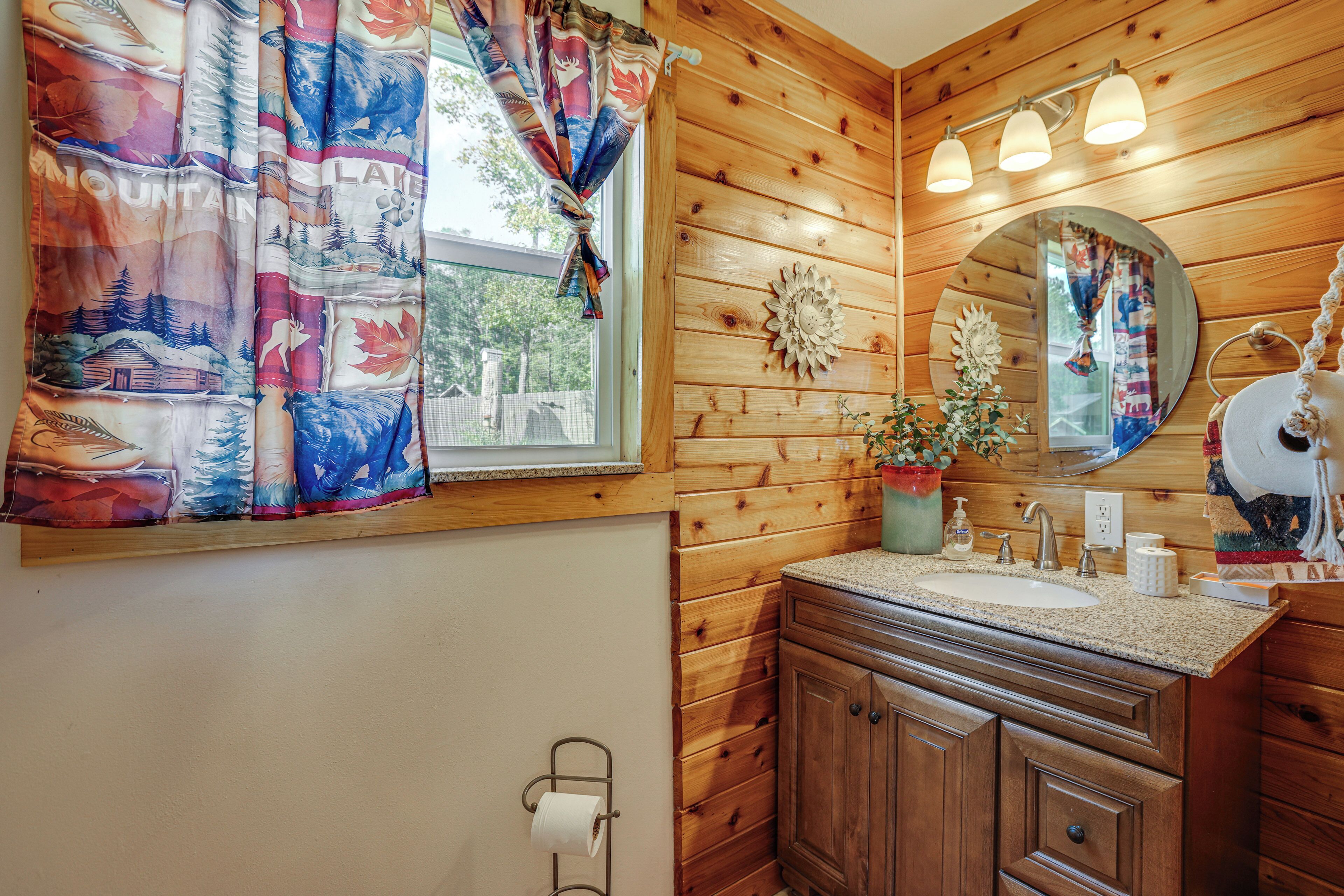 Ein Badezimmer einer Holzhütte mit einem Holzbadezimmertisch, einem Waschbecken, einem Spiegel und einem Fenster mit bunten Vorhängen.