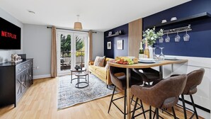 En moderne stue med sofa, spisebord med stole, fjernsyn og et køkkenområde med køleskab og vinglas.