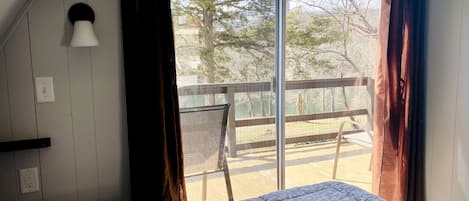 Una habitación con cama, ventana con cortinas, una silla y vistas a un área exterior con árboles y una cerca.