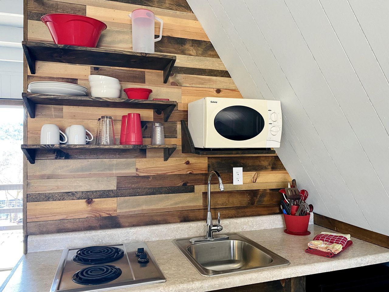 A kitchen with wooden shelves, a microwave, a sink, and a stovetop.
