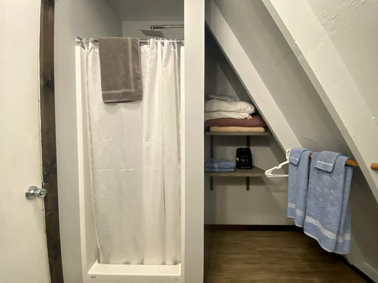 A bathroom with a shower curtain, a towel hanging on the door, a shelf with folded towels, and a staircase leading to an upper level.