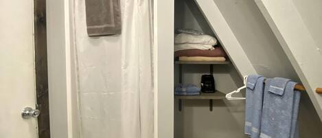 A bathroom with a shower curtain, a towel hanging on the door, a shelf with folded towels, and a staircase leading to an upper level.
