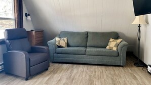 Ein Wohnzimmer mit grauem Sofa, einem schwarzen Sessel, einem Holztisch, einer Stehlampe und einem Fernseher.