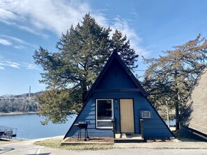 Une petite cabane triangulaire située au bord d’un lac, avec une porte et une fenêtre.