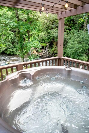 A hot tub with a wooden deck and railings, surrounded by lush greenery and a stream. - Group 07) Creekside Cove Yurt (Topton)