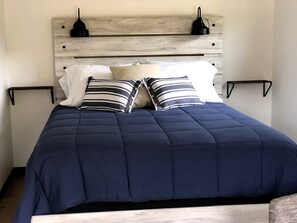 Un lit recouvert d’une courtepointe matelassée bleu marine, agrémenté de deux coussins à rayures et doté d’une tête de lit en bois sur laquelle sont fixées deux lampes murales noires.