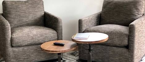 Dos sillones grises, una mesita redonda de madera con un control remoto y una alfombra estampada.