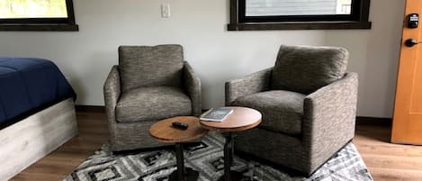 Una habitación con dos sillones grises, una pequeña mesa de madera, un tapete estampado y una cama con cubrecolcha azul.