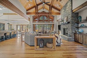 A spacious living area with a stone fireplace, wooden ceiling beams, and large windows overlooking a forest. - Meadowood Retreat with Access to Exclusive Golf (Sunriver)