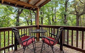 Eine Holzdielen-Terrasse mit Tisch und zwei Stühlen, umgeben von einem Geländer, mit Blick auf ein bewaldetes Gebiet.
