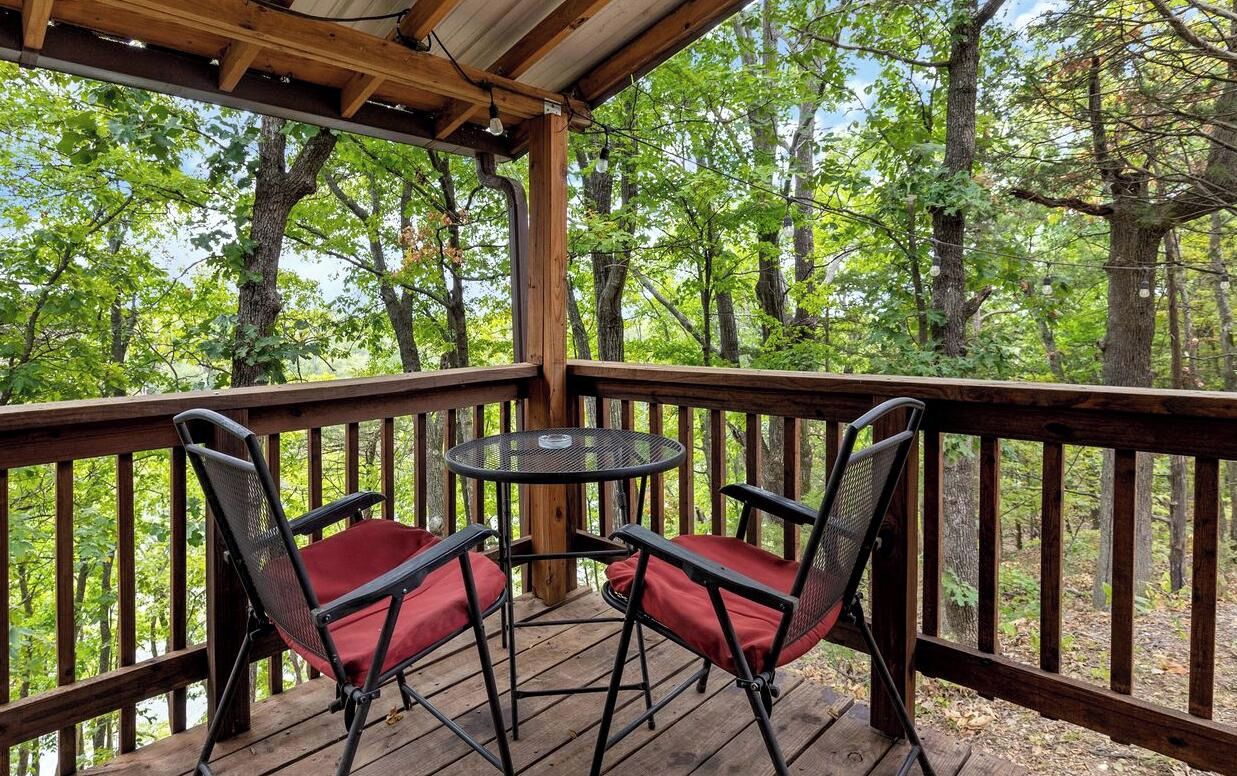 Una terraza de madera con una mesa y dos sillas, rodeada por una barandilla de madera, con vistas a un área boscosa.