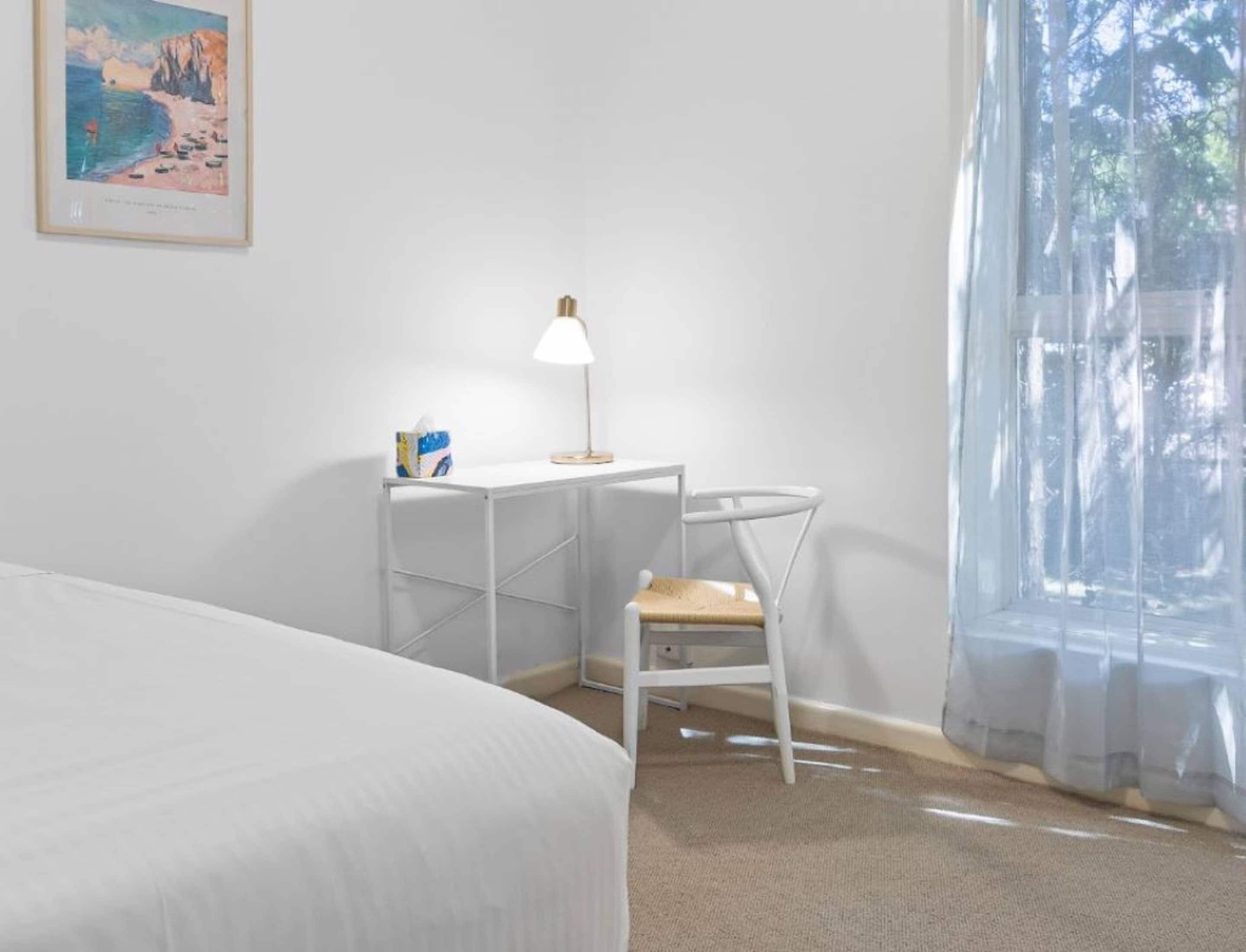 A bedroom with a bed, a desk with a chair, a lamp, and a window with sheer curtains.