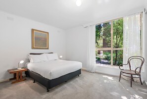A bedroom with a bed, a nightstand with a lamp, a chair, a window with curtains, and a painting on the wall. - Charming Inner-city Stay- Space, Style & Location (Melbourne)