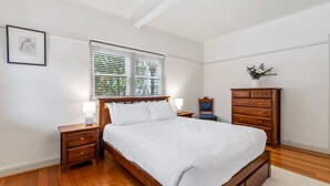 Een slaapkamer met een houten bed, nachtkastjes, een dressoir en een raam met jaloezieën.