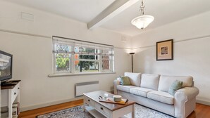 Een woonkamer met een witte bank, een salontafel, een kamerkleed met patroon, een raam met zonwering en een ingelijst kunstwerk aan de muur.