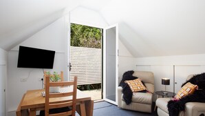 A room with a wooden dining table and chairs, a sofa with patterned cushions, a small side table with a lamp, and a television mounted on the wall. - Woolwich Studio Bliss, Private Oasis by the Water (Sydney)