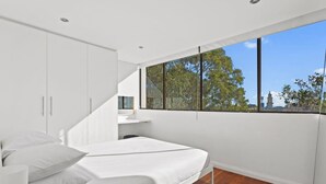 Une chambre moderne avec un lit, des armoires blanches et une grande fenêtre donnant sur des arbres et un immeuble.