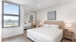 A modern bedroom with a large bed, bedside tables, a mirror, and a window. - 3-bed Apartment in Beautiful Kiama With Study Nook (Kiama)