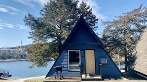 Una cabaña pequeña y triangular junto a un lago, con puerta y ventana.