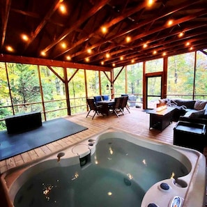 A hot tub in a covered outdoor area with wooden flooring, seating, and large windows. - Tiny Luxury Getaway: Cabin in Todd, North Carolina with Hot Tub, Scenic Views, and Lounge Perfection (Todd)