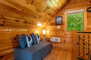 Een gezellig interieur van een chalet met een bank, kussens, een lamp en een raam met uitzicht op groen.