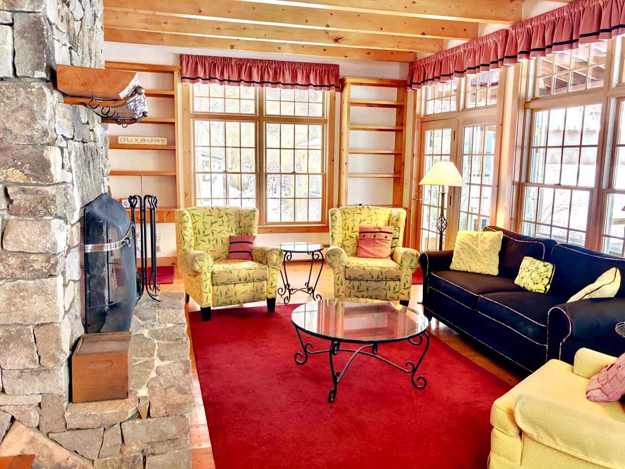 A cozy living room with a stone fireplace, yellow patterned armchairs, a blue sofa, and a red carpet.