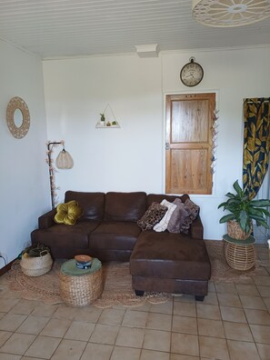 Ein Wohnzimmer mit einem braunen Ecksofa, einer Wanduhr, einer Holztür und einem Blumentopf.