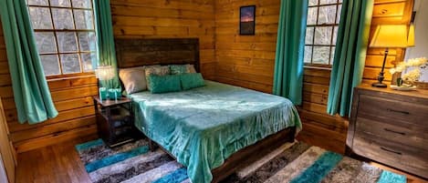 Une chambre dans une cabane en bois, avec un lit, une table de chevet, une commode et une fenêtre avec des rideaux.