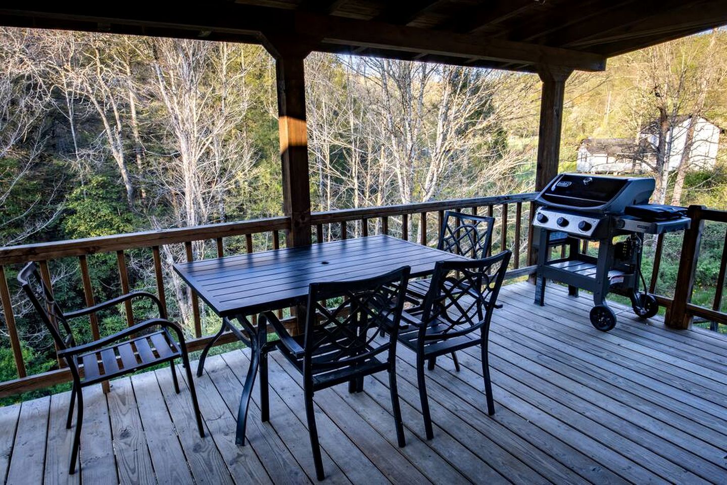 A covered outdoor deck with a table and chairs, a grill, and a view of trees and a house.