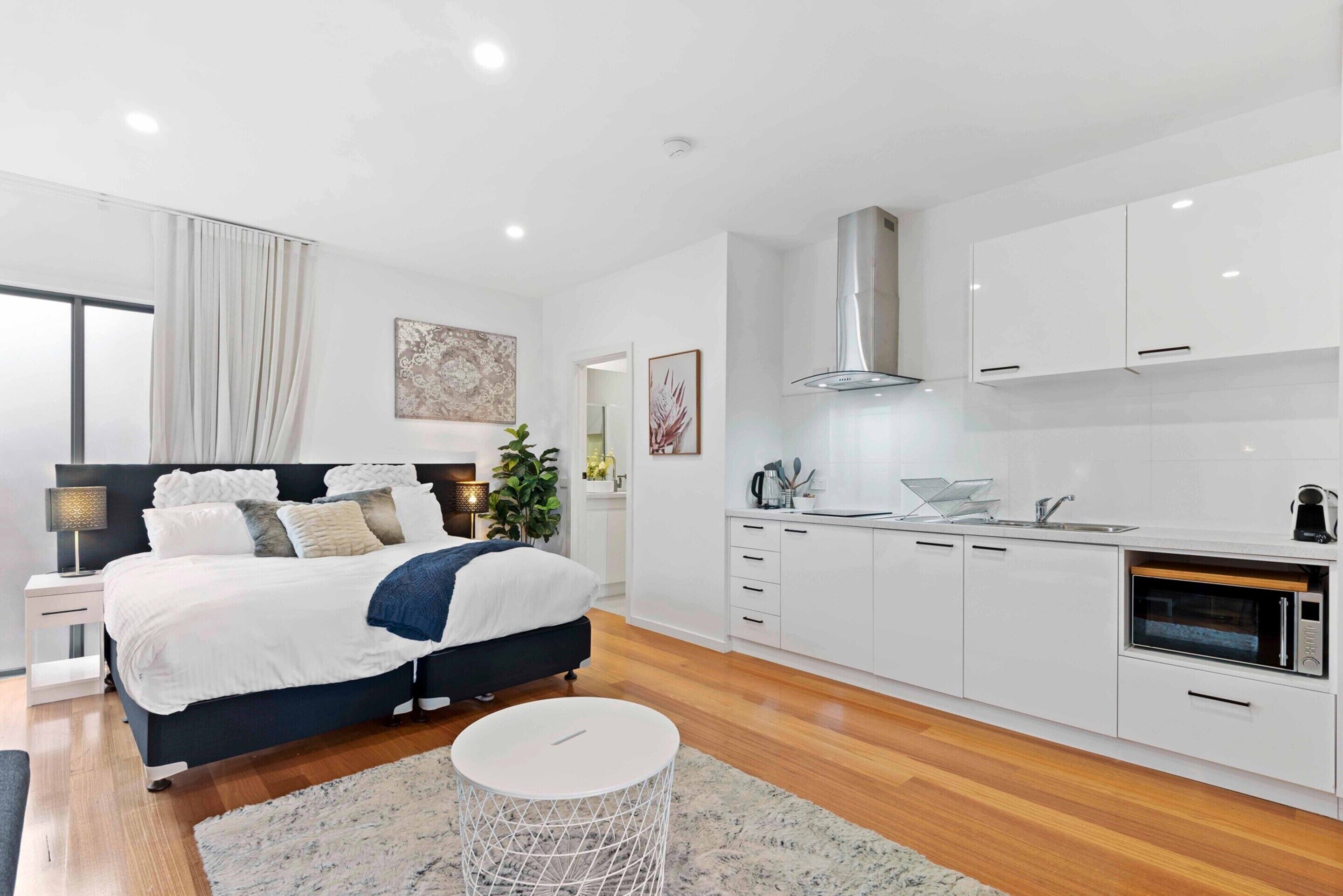 A modern bedroom with a large bed, bedside table, and a nightstand. There is a small round table in the center of the room. The room has white walls and a wooden floor.