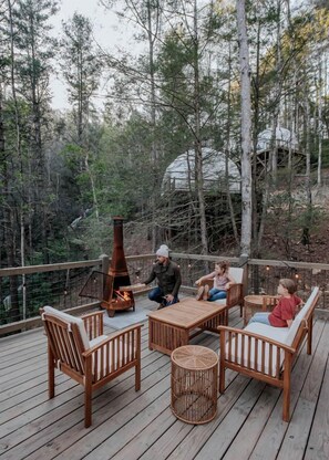 Eine Holzterrasse mit Gartenmöbeln, einer Feuerstelle und Blick auf einen bewaldeten Bereich.