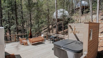 Een houten terras met buitenmeubilair, een jacuzzi en een koepelstructuur van glas, gelegen in een bosrijk gebied.