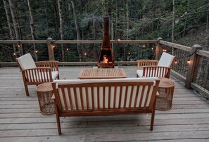 Una terraza de madera con un brasero en el centro, rodeada de sillones de mimbre y un banco de madera.