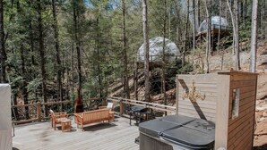 Una terraza de madera con mobiliario exterior, una bañera de hidromasaje y una estructura con cúpula de vidrio en un área boscosa.