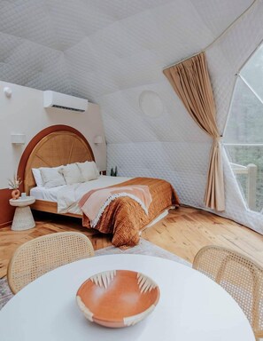 Un dormitorio con una cama, una mesa redonda, sillas de mimbre, una mesita de noche con un florero y una ventana con cortinas.