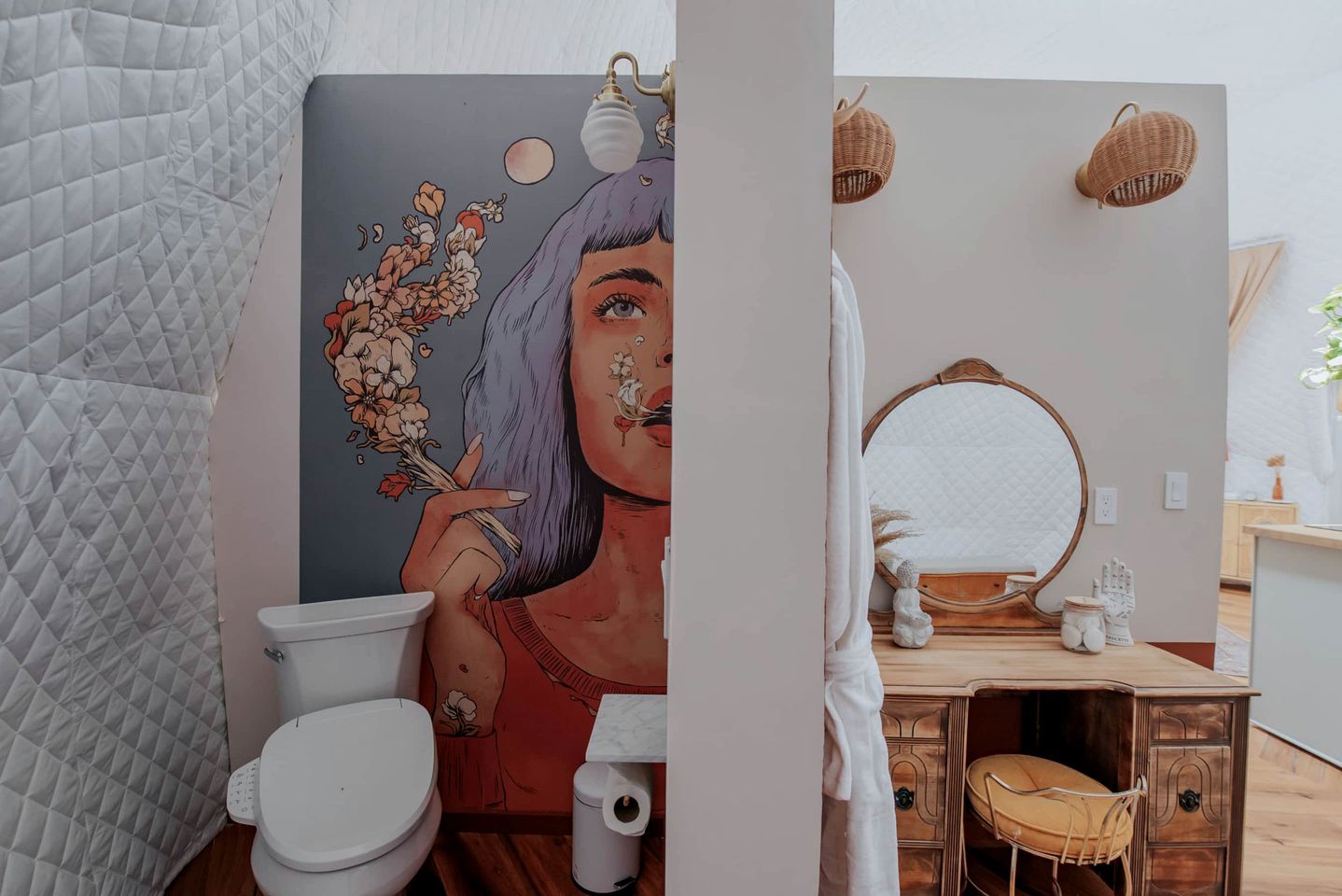 A bathroom with a toilet, a wall-mounted mirror, a wooden vanity, and a wicker lamp.