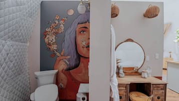 A bathroom with a toilet, a wall-mounted mirror, a wooden vanity, and a wicker lamp.