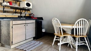 Una cocina con estantes de madera, un refrigerador, un microondas y una zona de comedor con sillas blancas y una mesa.