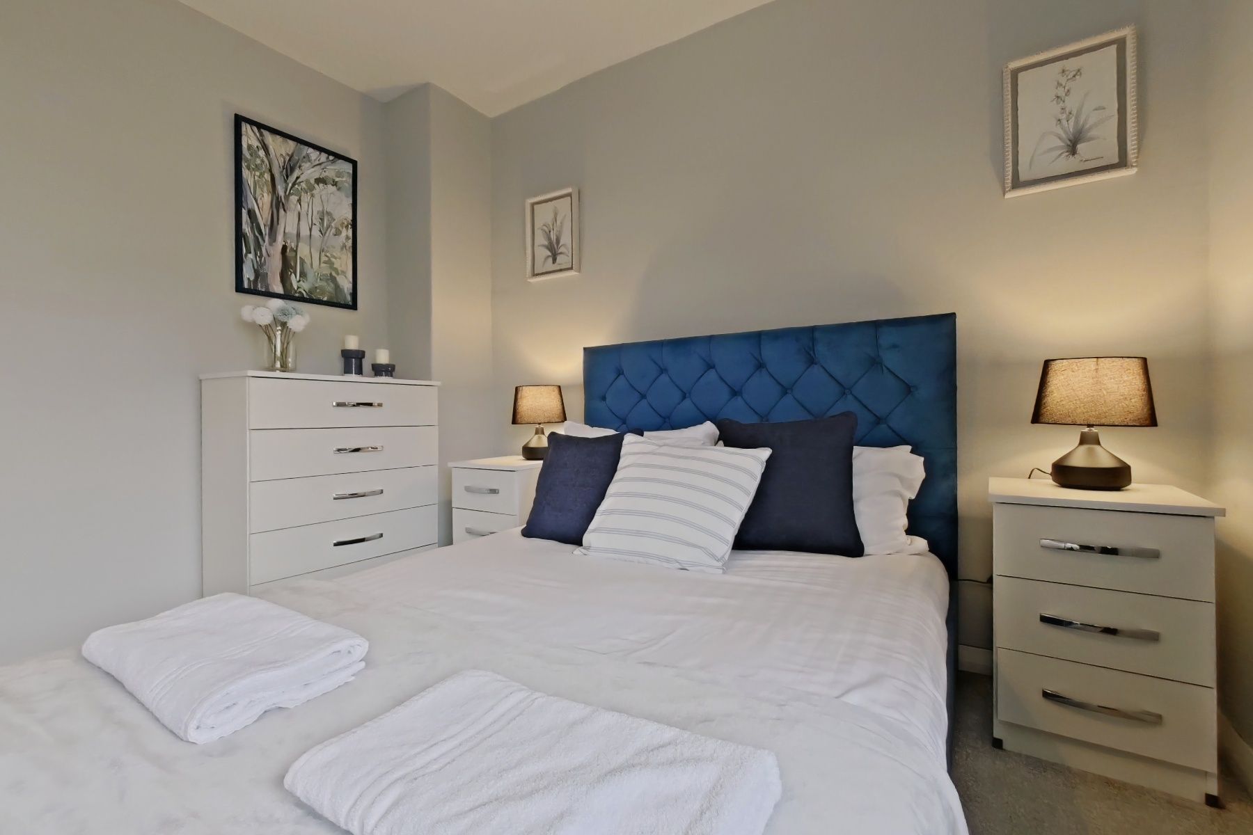 A bedroom with a large bed, two bedside tables with lamps, a dresser, and framed artwork on the walls.