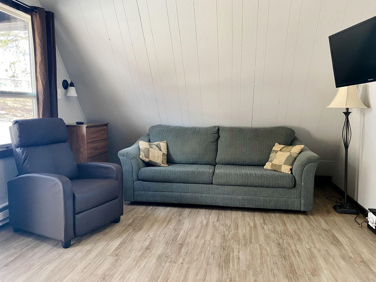 A living room with a grey sofa, a black armchair, a wooden side table, a floor lamp, and a TV.