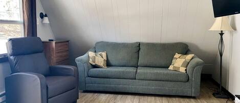 Un salon avec un canapé gris, un fauteuil noir, une table d’appoint en bois, un lampadaire et un téléviseur.