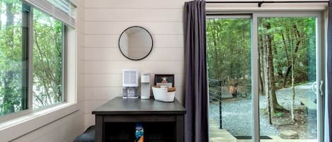 A room with a desk, a coffee maker, a basket, a mirror, and a view of a forested area through a glass door.