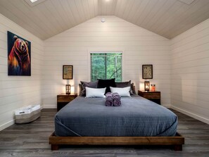Un dormitorio con techo inclinado, una cama grande con colcha azul, dos mesitas de noche con lámparas, una ventana con persianas y un cuadro de un oso en la pared.