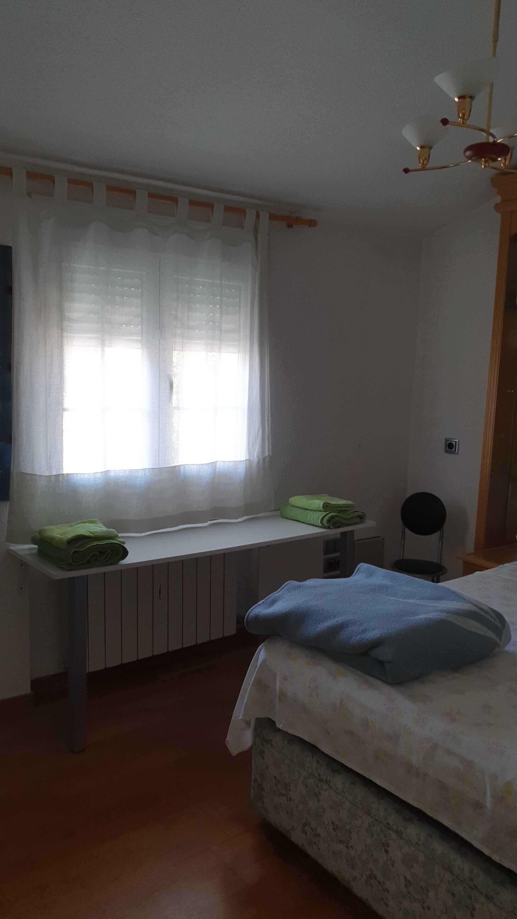 Ein Schlafzimmer mit Bett, Heizkörper, einem Fenster mit Vorhängen, einem kleinen Tisch mit Handtüchern und einem Stuhl.