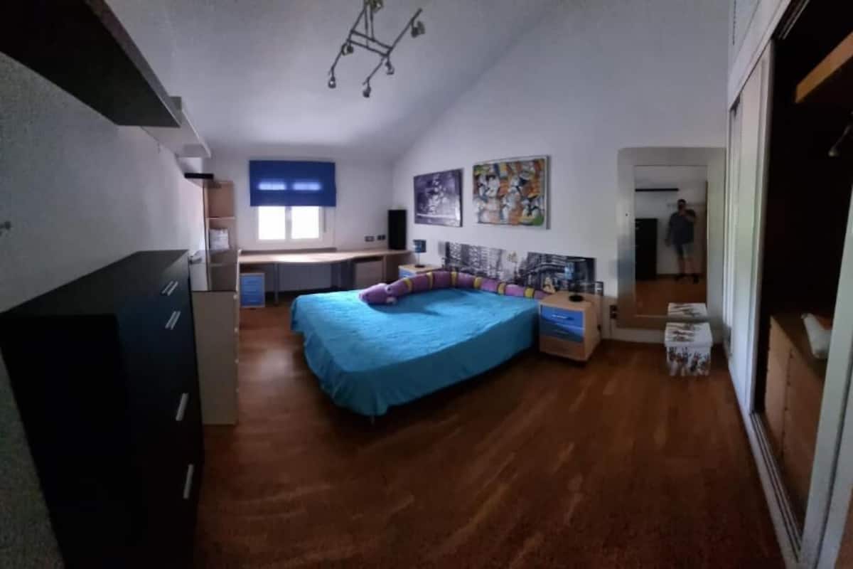Ein Schlafzimmer mit einem Bett, einem Schreibtisch und einem Stuhl. Es gibt ein Fenster mit Jalousien, eine Tür und eine Wand mit Kunstwerken.