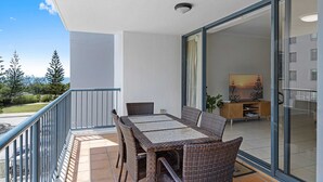 En balkon med et bord og stole, en skydedør der fører til en opholdsstue, samt udsigt til træer og en bygning i baggrunden.