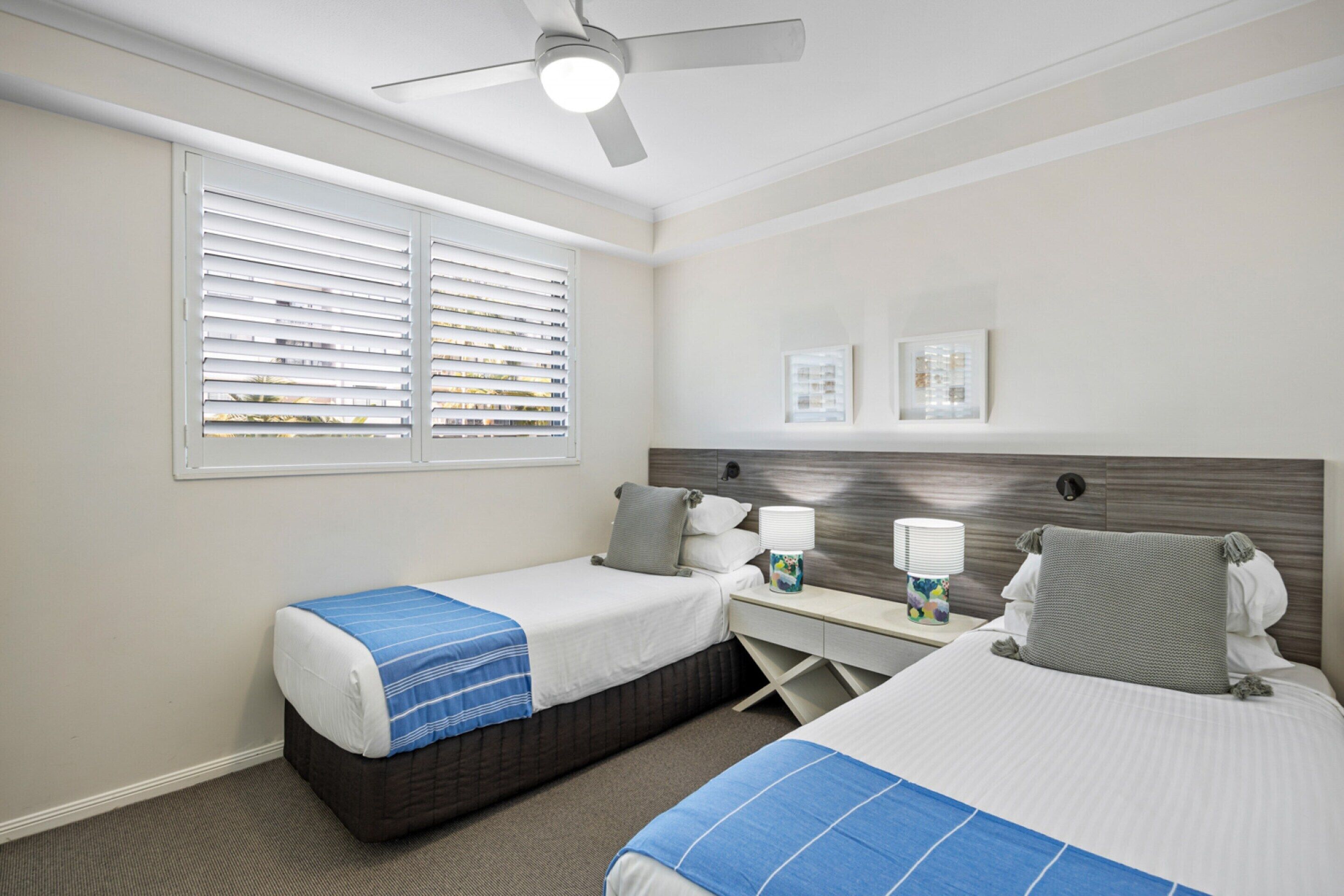 A hotel room with two beds, a desk, a chair, a ceiling fan, and a window with blinds.