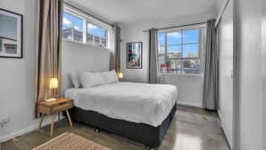 A modern bedroom with a large bed, bedside lamps, a view of the city through large windows, and a cityscape painting on the wall. - Bright 1-bed Unit With Balcony in Central Adelaide (Adelaide)