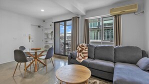 A modern living room with a grey sofa, a round wooden coffee table, and a glass dining table with chairs. There is a large window with curtains, an air conditioning unit, and a view of buildings outside. - Bright 1-bed Unit With Balcony in Central Adelaide (Adelaide)